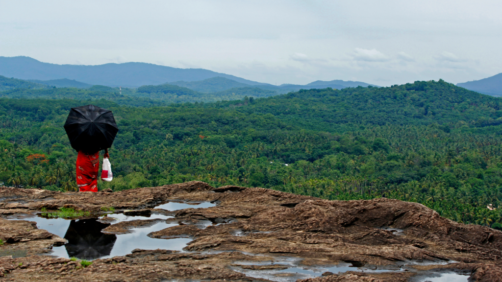 A woman in a red dress stands on a rocky hilltop, holding a black umbrella. She gazes out at a lush green valley with mountains in the distance. Monsoon Rain has created small puddles on the rocky surface.