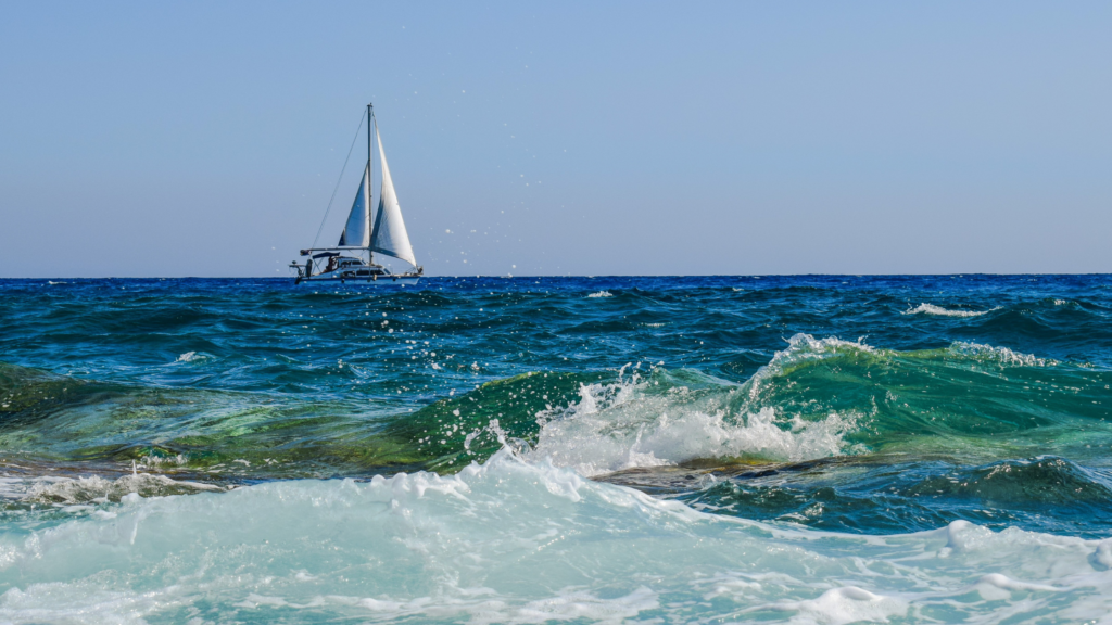 A sailboat with white sails glides across a blue ocean. Waves crash against rocky shore in the foreground, creating white foam.