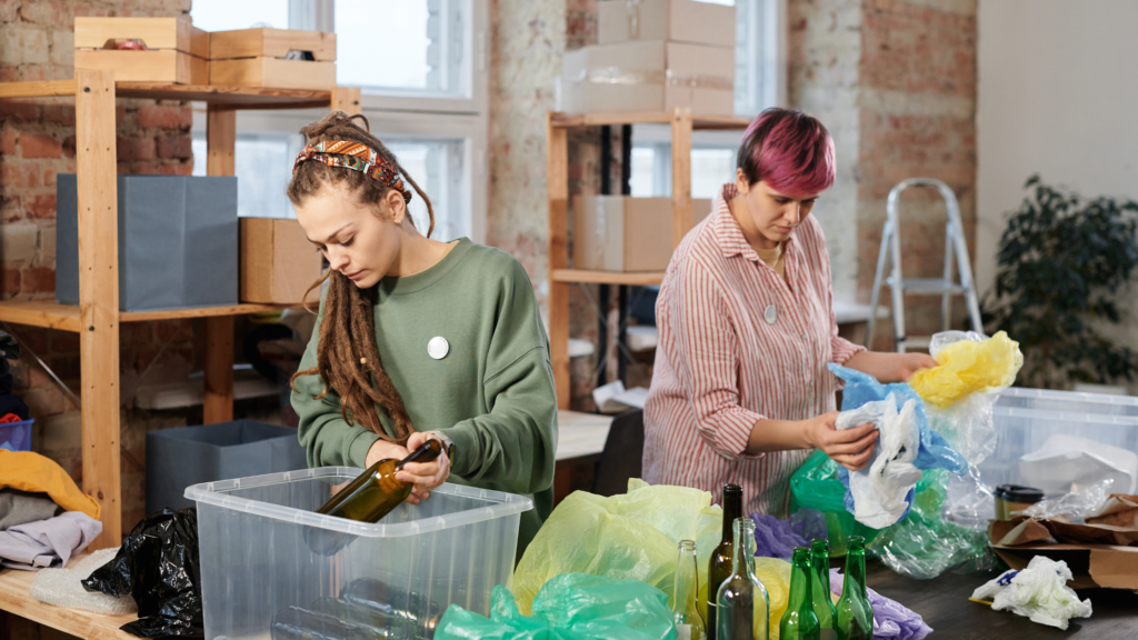 Two individuals in a workshop-like room, sorting recyclable materials into bins. One person with dreadlocks is sorting textiles and glass bottles, while another with pink hair is sorting plastic bags. The room is filled with various recyclable materials and has a casual, functional atmosphere.