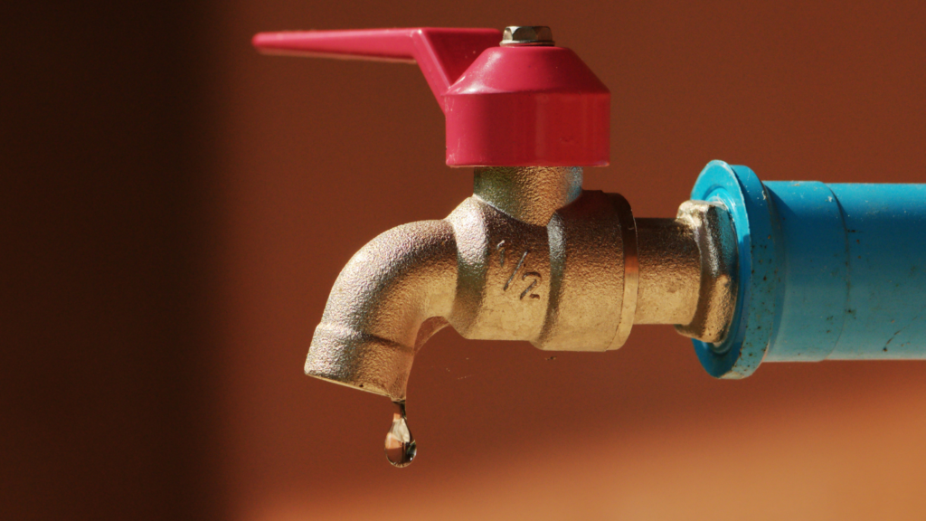 close-up of a brass water tap with a red handle, connected to a blue PVC pipe, with a single water droplet hanging from the spout, suggesting a slight leak. The background is out of focus with a warm brown tone, highlighting the issue of water wastage through leaky faucets.