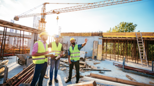 Three construction workers in hard hats and safety vests stand at a building site, discussing plans against a backdrop of a crane and a partially constructed building.