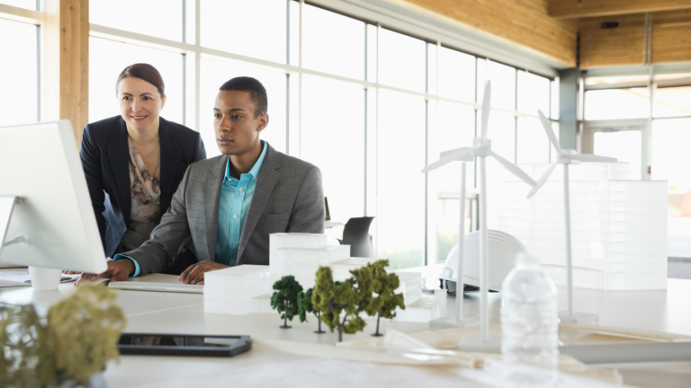 Two architects, one male and one female, review blueprints on a computer in a modern office. The foreground features a model of a sustainable building project with miniature wind turbines and trees. Company representing circular economy examples