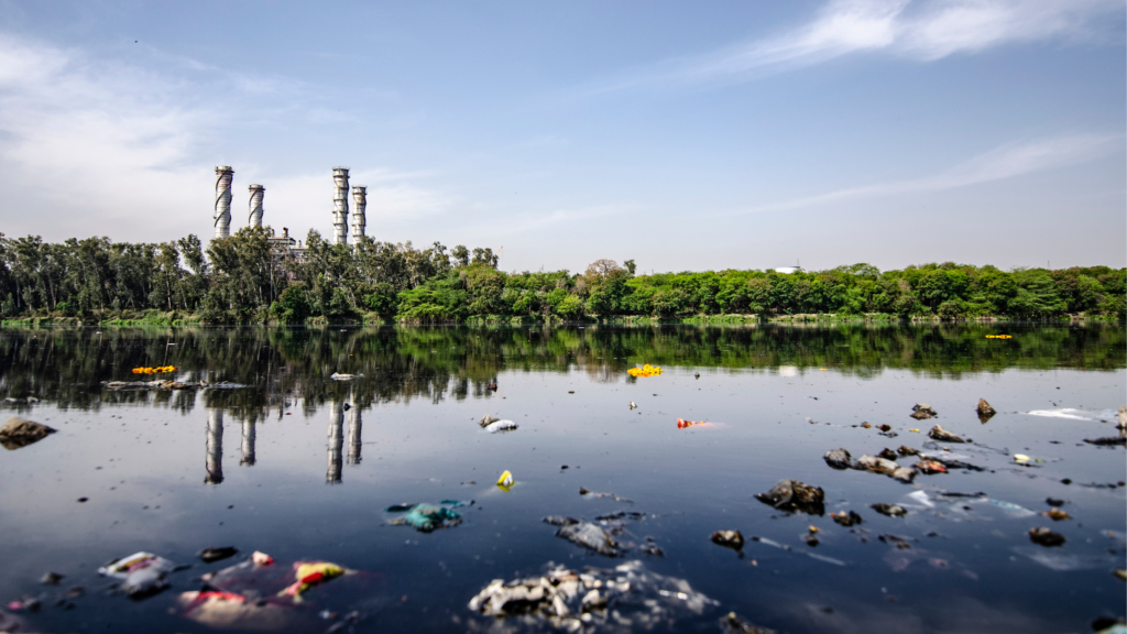 A polluted lake with plastic waste and debris floating on the surface. Industrial chimneys are visible in the background.