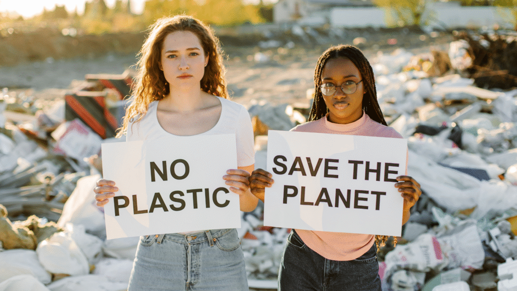 Two young women standing in front of a large pile of trash or plastic waste. One woman holds a sign that reads 'NO PLASTIC'. The other woman holds a sign that reads 'SAVE THE PLANET'. Depicting International day of Environment 2025 on plastic pollution